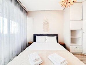 A neatly arranged hotel room with a bed, white curtains, a wall hanging, and a wooden nightstand. - Bright Modern 1BR · 4-min Walk to the Sea · Alimos Riviera (Alimos)