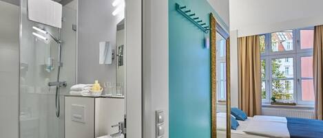 Una habitación de hotel moderna con cabina de ducha de vidrio, lavabo, cama con manta azul y ventana con cortinas.