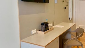A modern hotel room with a flat-screen TV mounted on the wall, a small desk with a coffee maker, a chair, and a door with a round mirror. - Hotel 38  (Berlin)