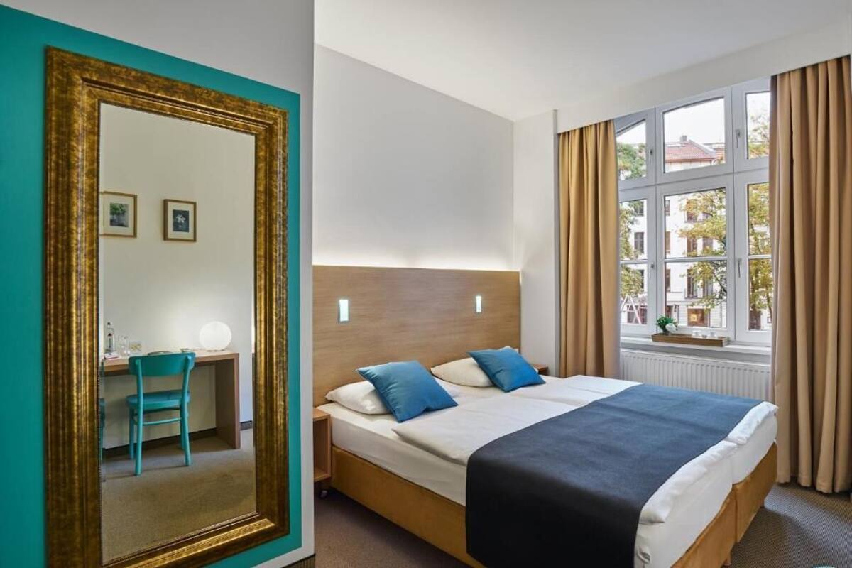 Habitación de hotel con cama, escritorio con silla, espejo grande y ventana con cortinas.
