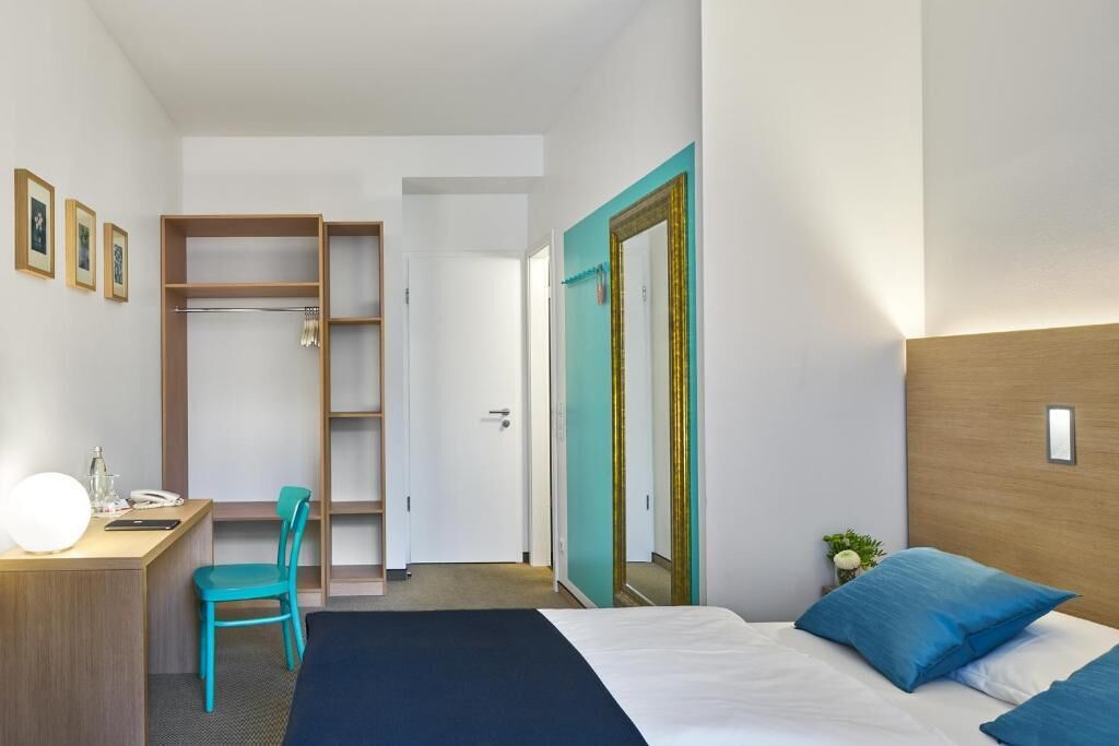 A hotel room with a bed, a desk with a chair, a wardrobe, and a mirror.