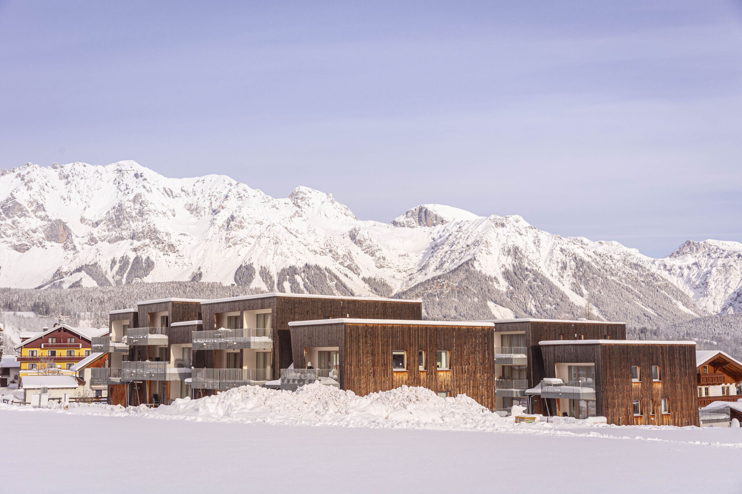 Ein modernes Gebäude mit Balkonen, einer Holzfassade und einem Bergpanorama im Hintergrund.