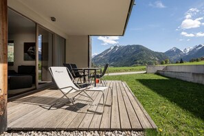 Eine Holzterrasse mit Gartenmöbeln, eine Glastür, die zu einem Raum führt, und Blick auf die Berge.