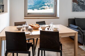 A dining area with a table set for two, chairs, a window with a mountain view, and a couch in the background. - Premium apartment #6.2  with 1 bedroom and sauna area (Rohrmoos-Untertal)