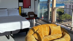 Een hotelkamer met een bed, een stoel, een nachtkastje en een groot raam met uitzicht op zee en bergen.