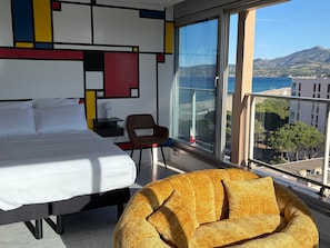 Ein Hotelzimmer mit Bett, Sessel, Nachttisch und einem großen Fenster mit Blick auf das Meer und die Berge.