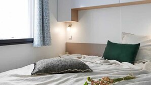 Ein Schlafzimmer mit Bett, Kissen, Nachttisch, Vorhängen an einem Fenster und einer Pflanze auf dem Fensterbrett.