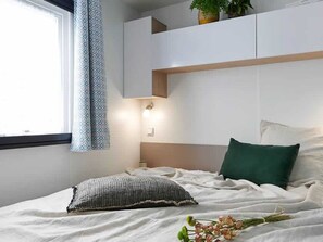 Ein Schlafzimmer mit Bett, Kissen, Nachttisch, Vorhängen an einem Fenster und einer Pflanze auf dem Fensterbrett.