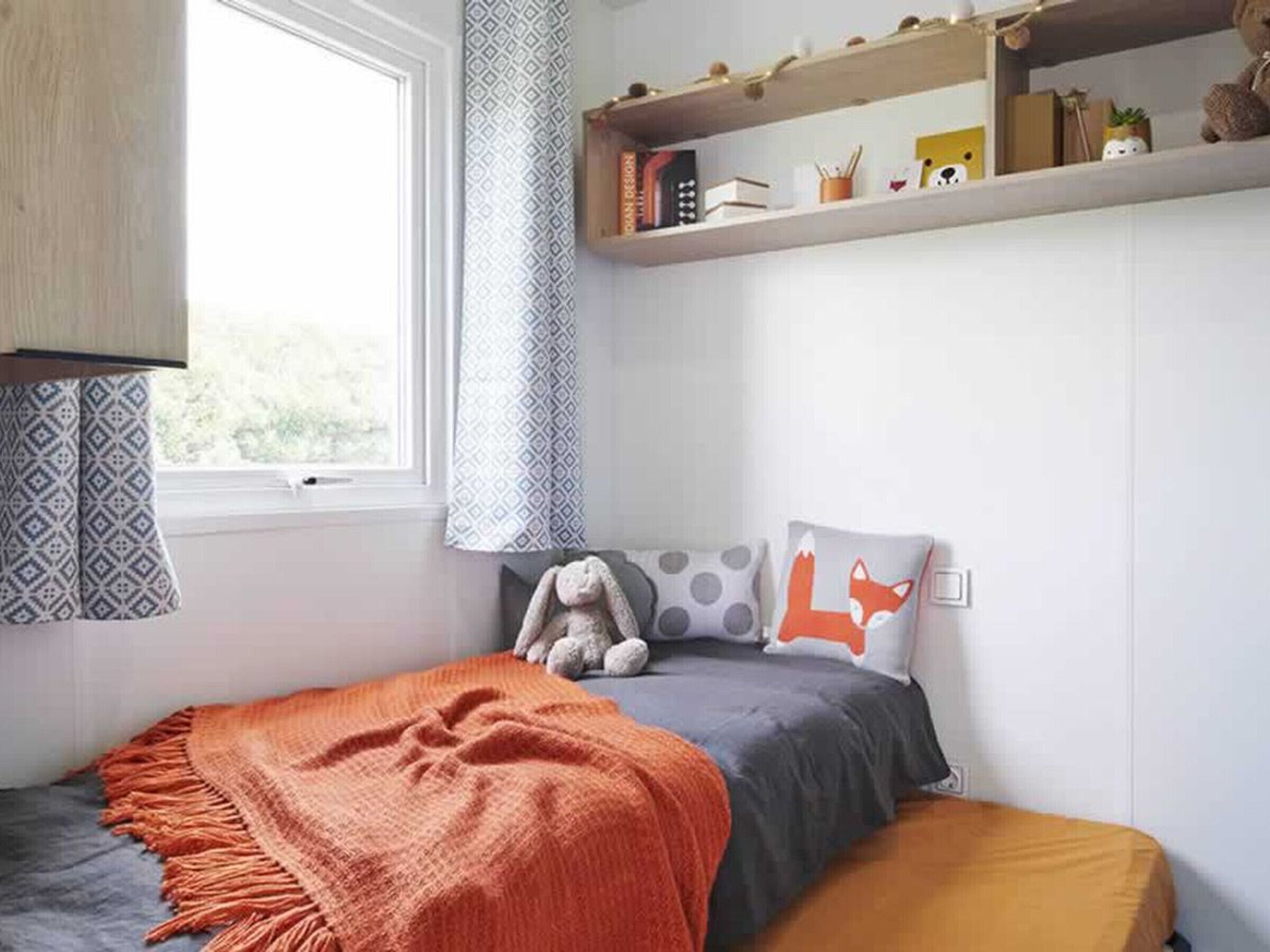 Une chambre à coucher avec un lit, une fenêtre, une étagère avec des livres et des objets décoratifs, et une couverture.