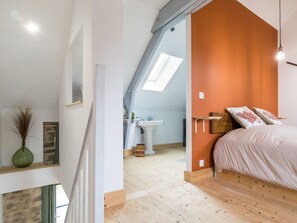 A bedroom with a sloped ceiling, a bed, a bedside table, a nightstand, a window, and a bathroom with a sink and mirror. - Charming cottage in Clis - 2 guests (Guérande)