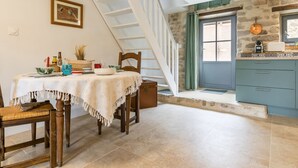 A dining area with a table set for two, a wooden chair, a staircase, a kitchen with blue cabinets, and a stone wall. - Charming cottage in Clis - 2 guests (Guérande)