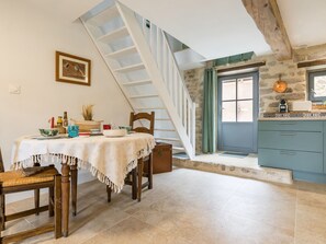 A dining area with a table set for two, a wooden chair, a staircase, a kitchen with blue cabinets, and a stone wall. - Charming cottage in Clis - 2 guests (Guérande)