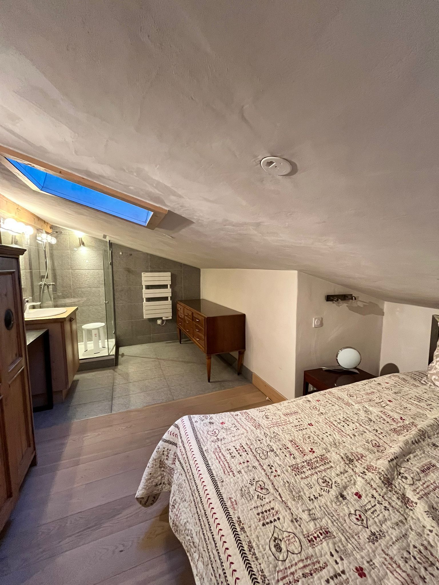Een slaapkamer met dakraam, een bed met beddengoed met patroon, een houten dressoir en een badkamer met douche en handdoekhouder.