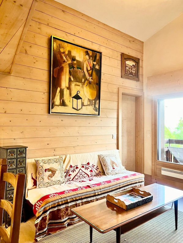 A cozy room with a wooden ceiling, a large painting, a sofa with patterned cushions, a wooden coffee table, and a window with a view. - Charming apartment facing Mont Blanc (combloux)