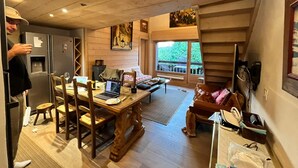 A cozy room with a wooden dining table, chairs, a laptop, and a sofa. There's a window with a view of greenery outside. - Charming apartment facing Mont Blanc (combloux)