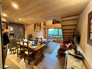 Une pièce chaleureuse avec une table à manger en bois, des chaises, un ordinateur portable et un canapé. Une fenêtre offre une vue sur la verdure environnante.