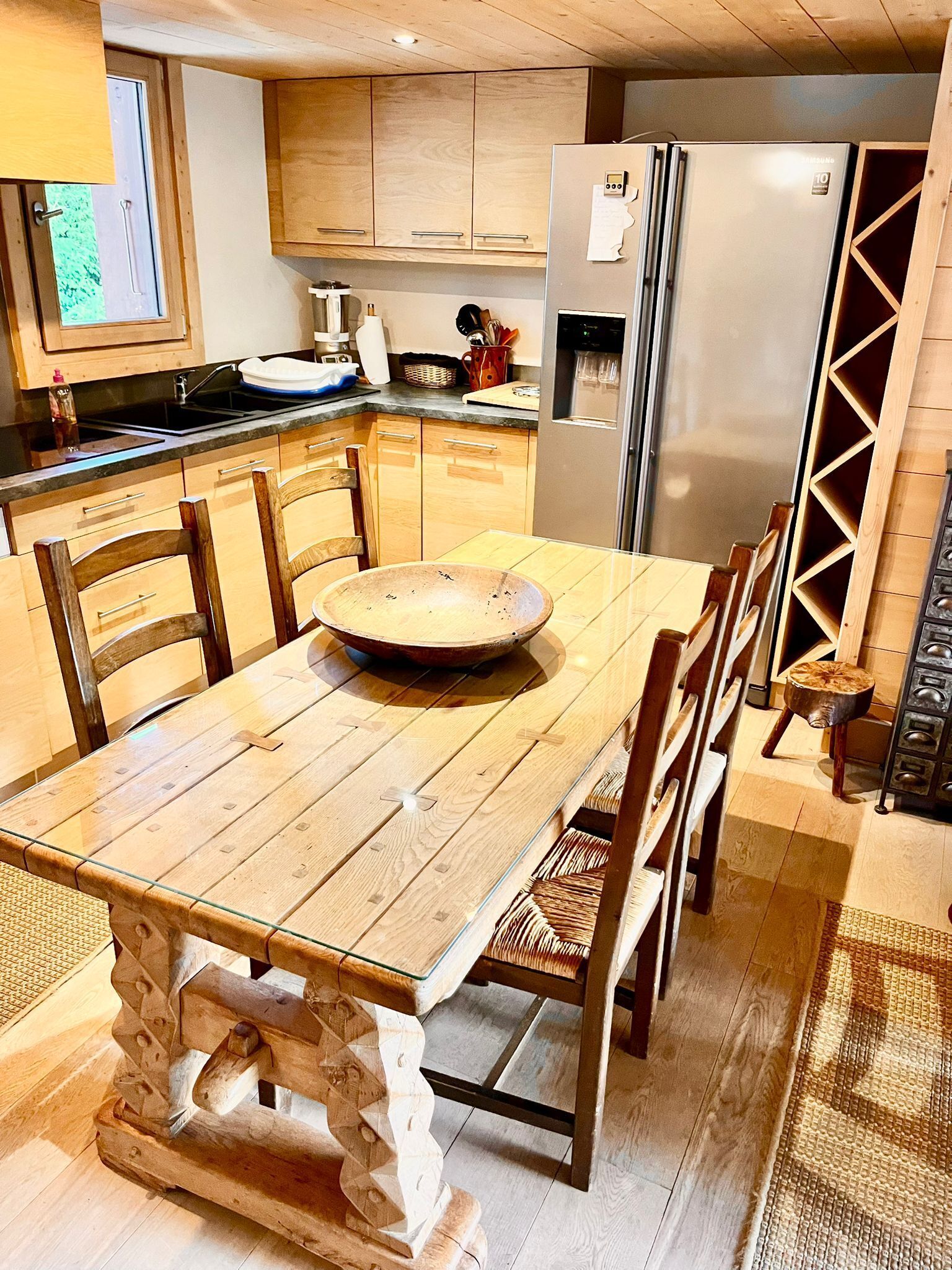 Une table de salle à manger en bois, avec des chaises, un grand saladier et un réfrigérateur, dans une cuisine dotée de meubles en bois et d’une fenêtre.