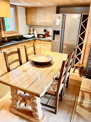 A wooden dining table with chairs, a large bowl, and a refrigerator in a kitchen with wooden cabinets and a window. - Charming apartment facing Mont Blanc (combloux)