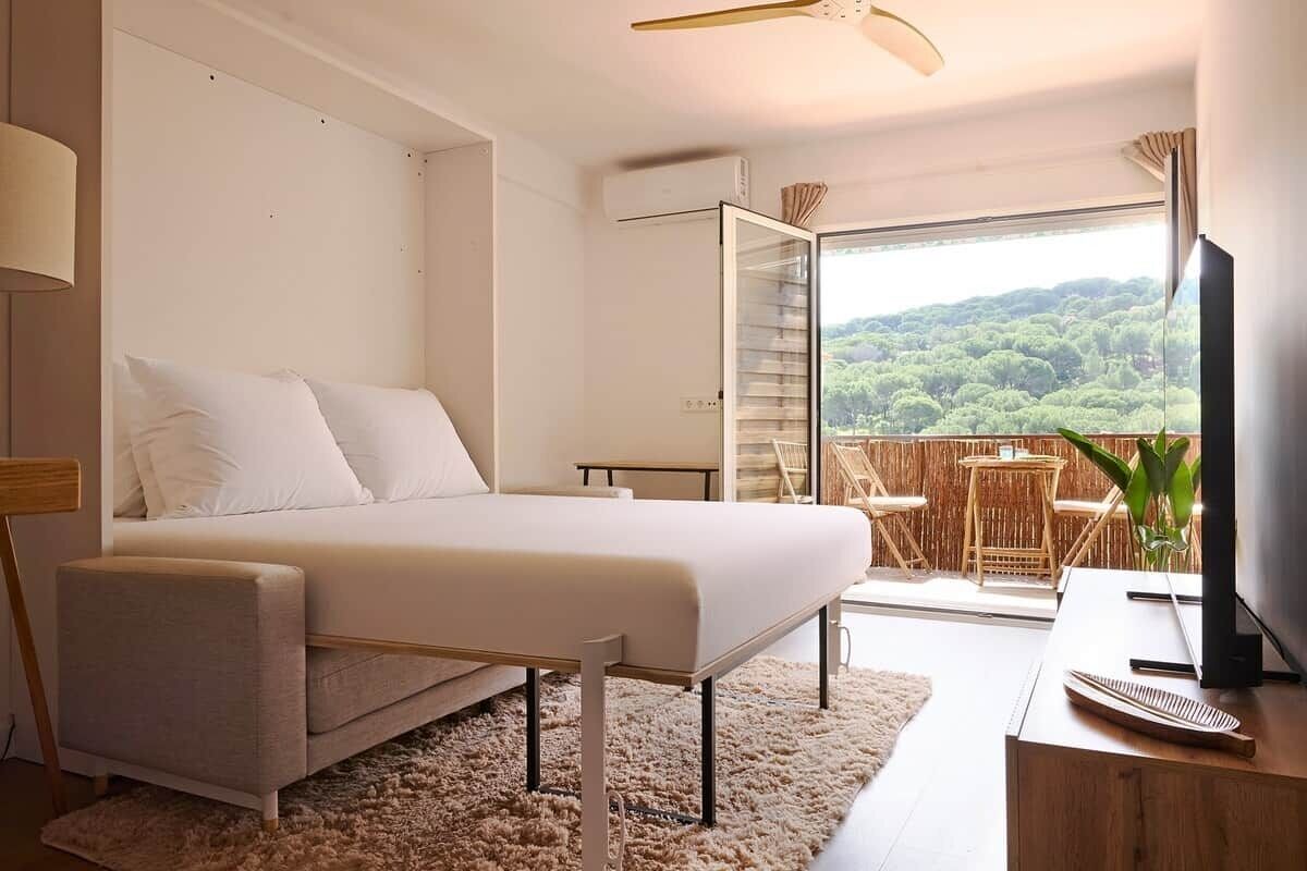 Ein modernes Schlafzimmer mit Bett, Schreibtisch, Fernseher und einem Balkon mit Blick auf Grünflächen.