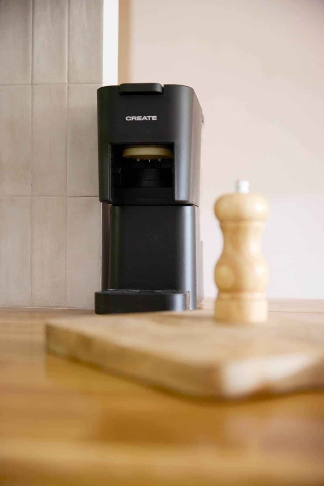 A black coffee machine with the word 'CREATE' on it, placed on a wooden board next to a wooden pepper grinder.