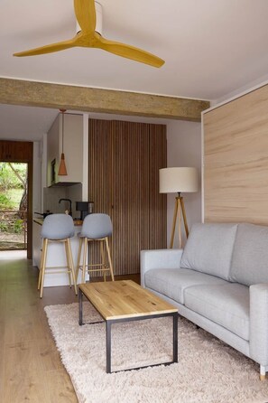 A modern living room with a grey sofa, wooden coffee table, two bar stools, a ceiling fan, and a kitchen visible in the background. - La Escondida Rural (San Martín de Valdeiglesias)
