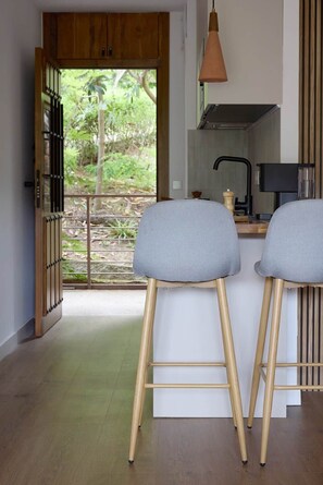 A kitchen with a view of greenery, two bar stools, a hanging pendant light, and a wooden cabinet. - La Escondida Rural (San Martín de Valdeiglesias)