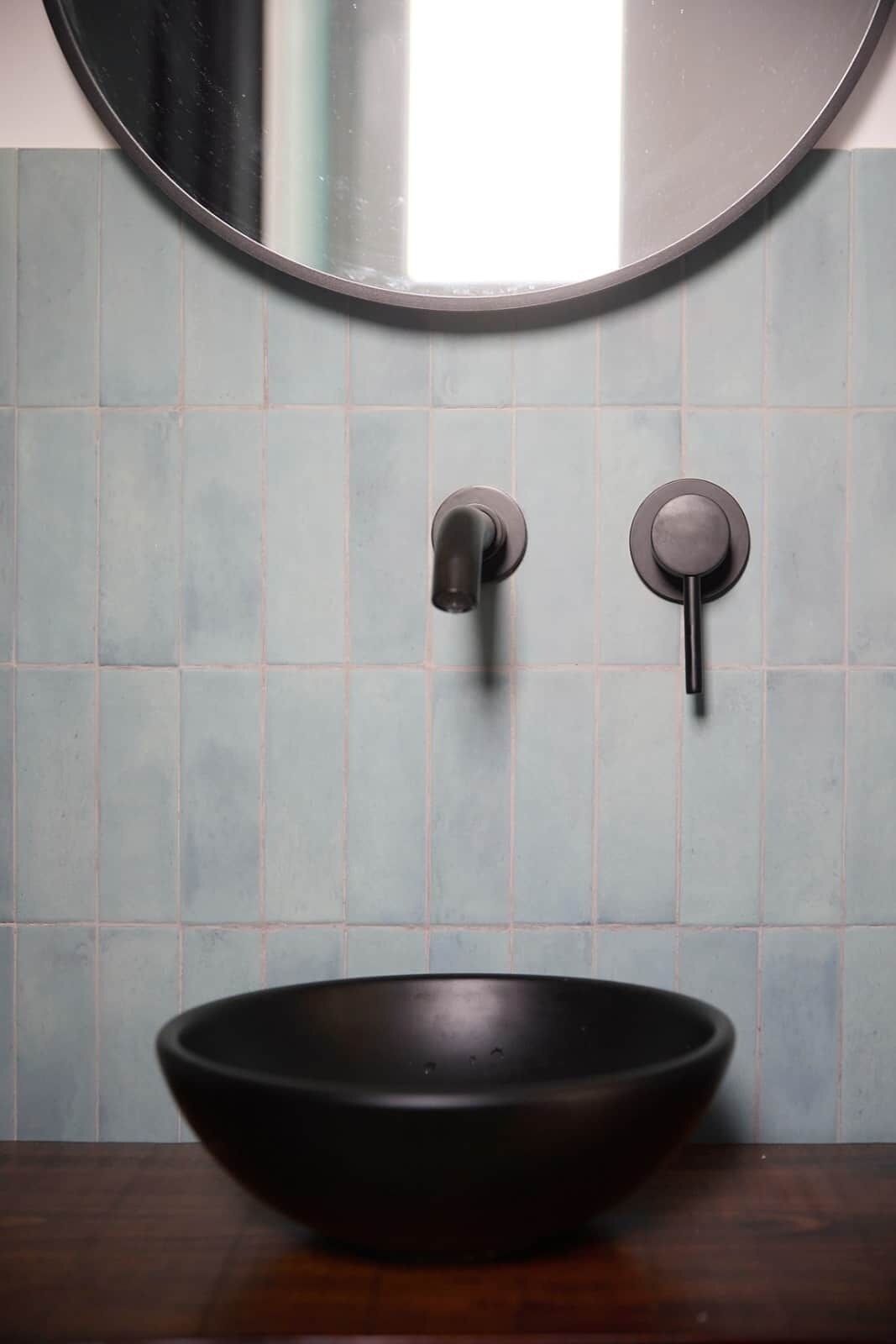 Un baño con un espejo redondo, un lavamanos tipo cuenco negro y dos grifos empotrados en la pared.