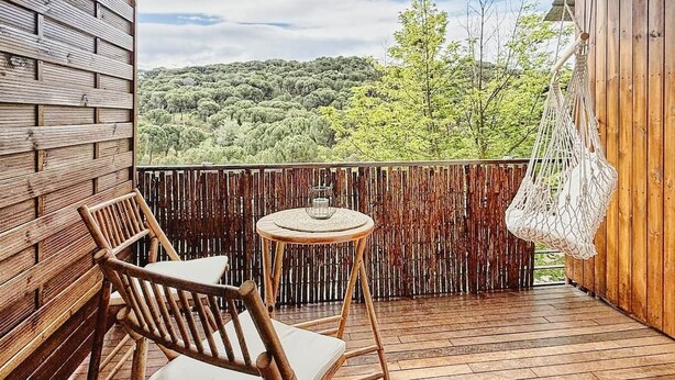 A wooden deck with a chair and table, a hanging macrame chair, and a view of a forested hill. - La Escondida Apartment at the San Juan B Reservoir (San Martín de Valdeiglesias)