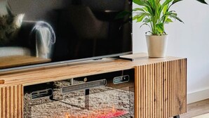 A modern living room with a flat-screen TV on a wooden stand, a potted plant, and a fireplace below the TV. - La Escondida Apartment at the San Juan B Reservoir (San Martín de Valdeiglesias)