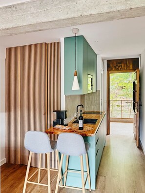 A modern kitchen with a wooden bar, two bar stools, a hanging pendant light, and a view of greenery outside. - La Escondida Apartment at the San Juan B Reservoir (San Martín de Valdeiglesias)