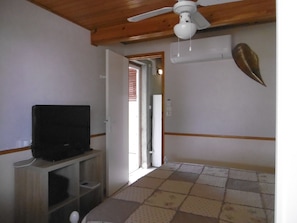 Ein Zimmer mit Deckenventilator, Fernseher auf einem Regal, einem gemusterten Teppich und einer Tür zu einem weiteren Raum.