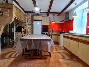 A dining area with a table, chairs, a stone wall, a kitchen with red walls, and a window. - Charming country cottage near Verdun (11 guests max) (Anoux)