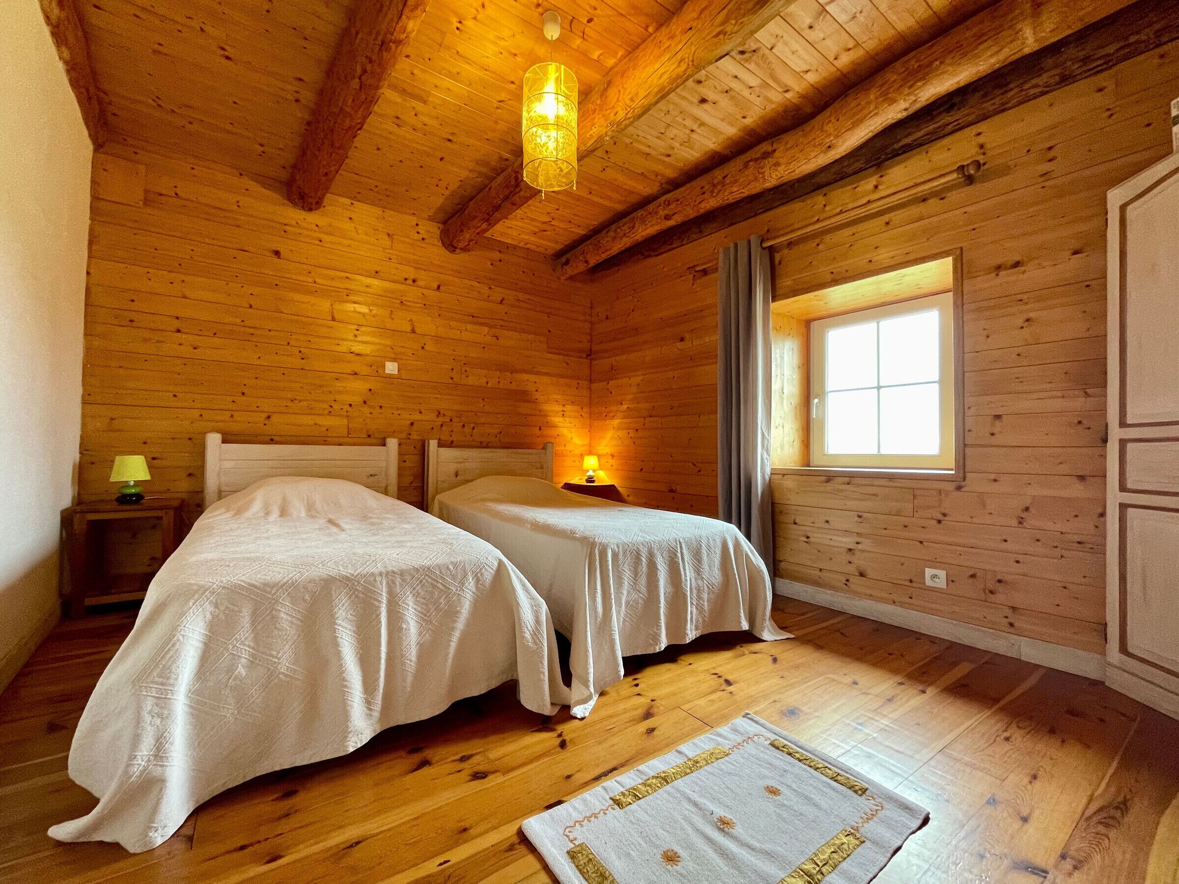 A wooden cabin room with two beds, a window, and a hanging light fixture.