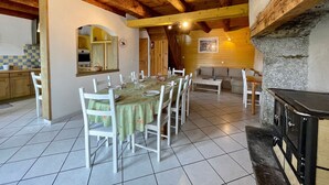 A dining area with a long table set for six, surrounded by white chairs, a stone fireplace, and a kitchen area with wooden cabinets and a tiled backsplash.