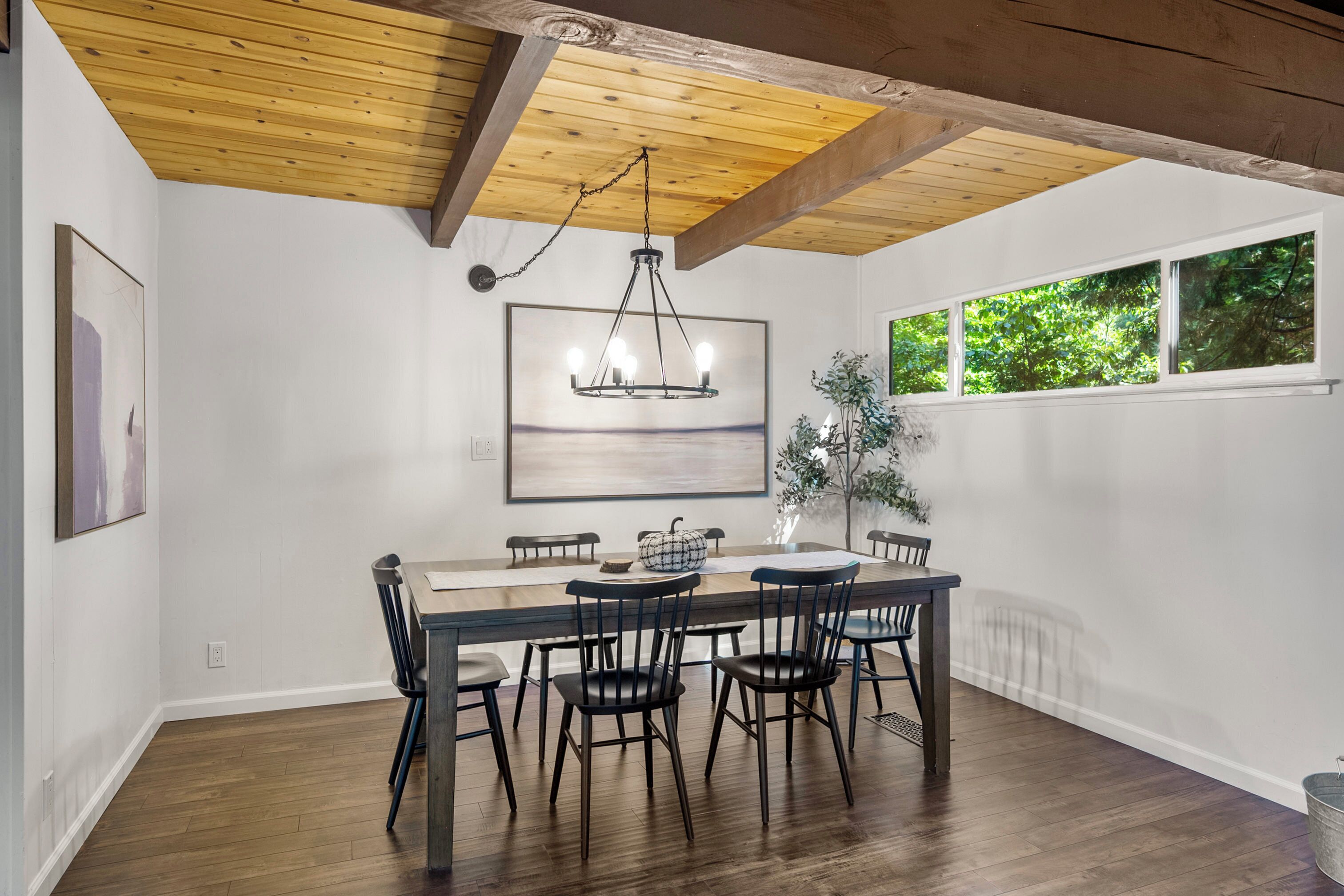 A dining area with a wooden ceiling, a chandelier, a large window, and a framed artwork on the wall.