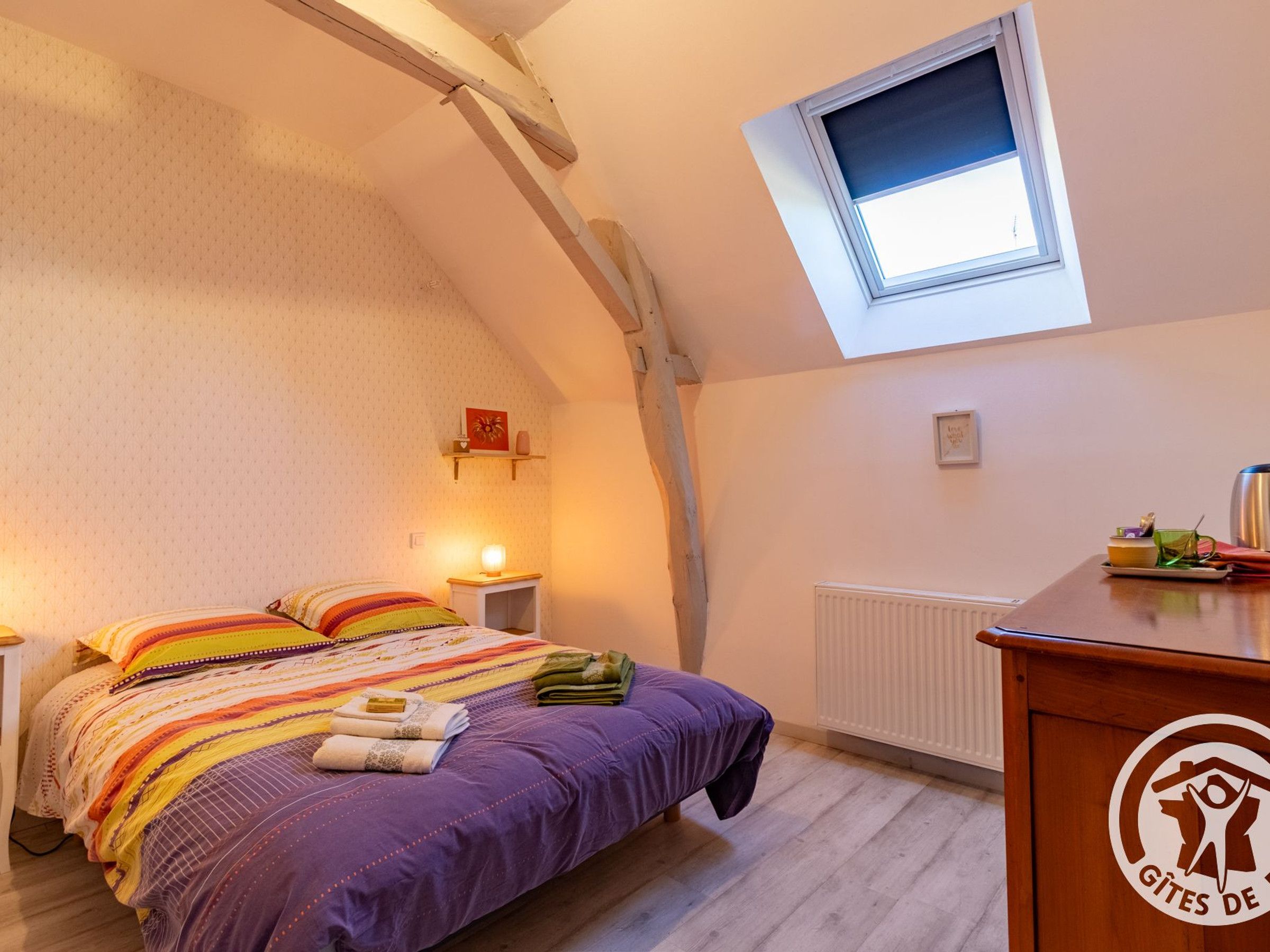 Une chambre avec un plafond en pente, un puits de lumière, un lit avec une literie colorée, une table de chevet avec une lampe et un radiateur.
