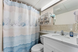 Et badeværelse med bruseforhæng, toilet, vask med spejl og en hårtørrer.