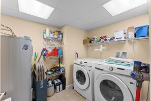 A laundry room with a washing machine, dryer, shelves with cleaning supplies, and a water heater. - Residence 207 at The Grand (Wildwood)