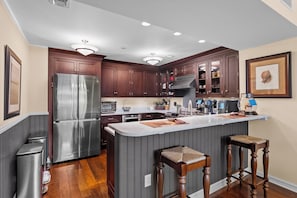 A modern kitchen with a stainless steel refrigerator, wooden cabinets, a central island with stools, and recessed lighting. - Residence 207 at The Grand (Wildwood)