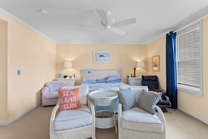 A bedroom with a bed, two chairs, a small table, a ceiling fan, and a window with curtains. - Residence 207 at The Grand (Wildwood)