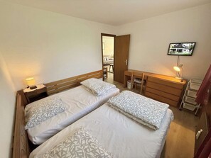 Ein Zimmer mit zwei Betten, einem Schreibtisch, einem Sessel, einem Fernseher und einer Tür zu einem anderen Raum.
