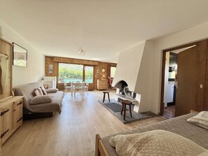 A spacious room with a sofa, a dining table, a fireplace, and a view of greenery outside. - Practical apartment with parking - 6 guests (Praz-sur-Arly)