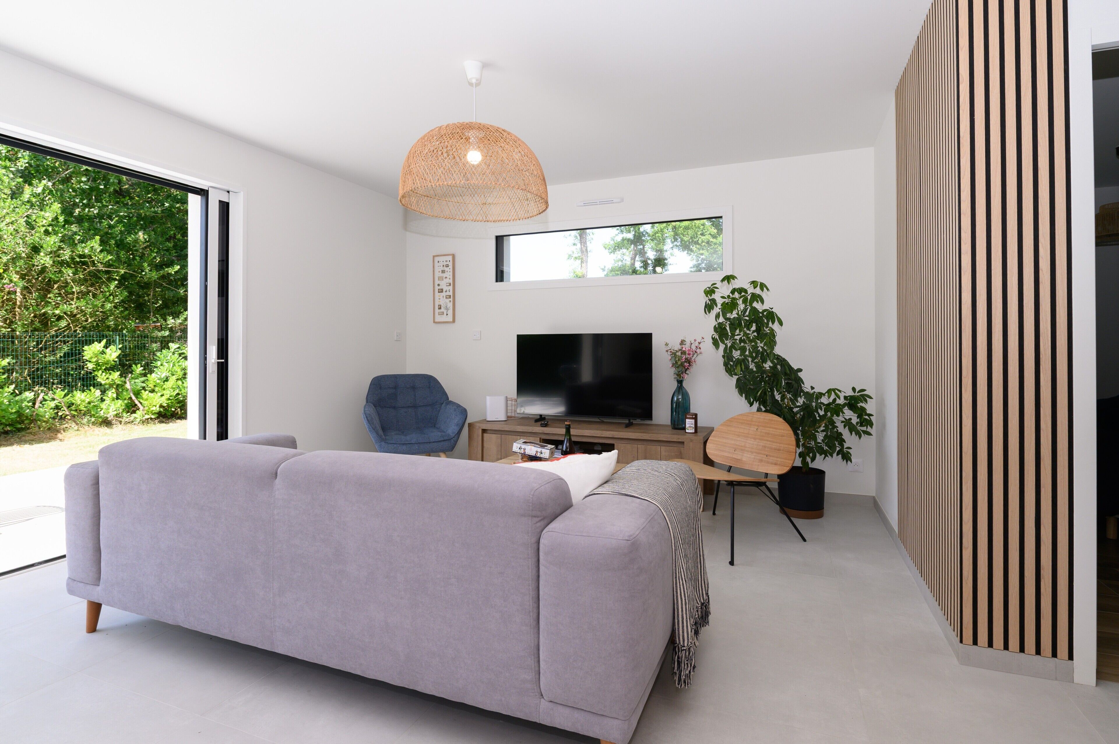 Una sala de estar moderna con un sofá gris, un televisor, una lámpara colgante de mimbre, un aparador de madera y un ventanal grande.