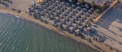 An aerial view of a coastal area with a beach, a row of modern houses, and a small resort area with umbrellas and lounge chairs.
