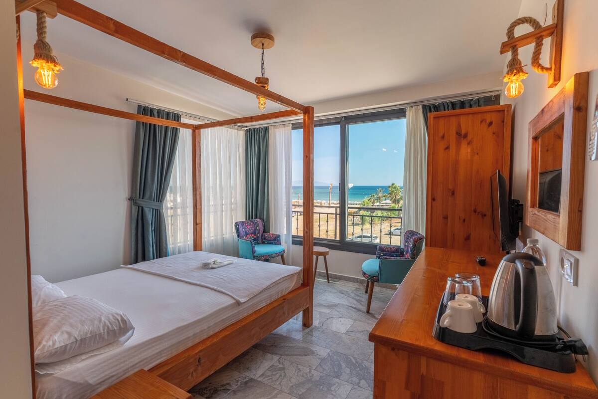 A room with a wooden four-poster bed, a small desk with a kettle and cups, and a view of the sea through large windows.