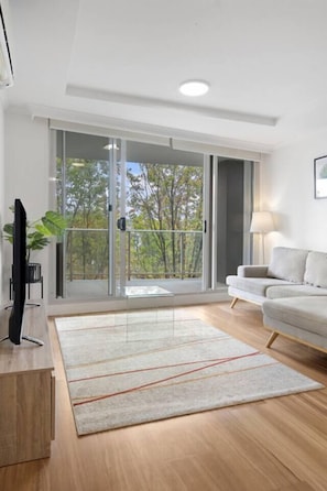 A modern living room with a flat-screen TV, a sofa, a lamp, and a large sliding glass door leading to a balcony. - Heart of city/modern 2BR/free parking/ANU/pool/gym (Braddon)
