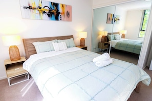 A neatly made bed with a quilted cover, two bedside lamps, a large mirror, and a painting on the wall. - Heart of city/modern 2BR/free parking/ANU/pool/gym (Braddon)