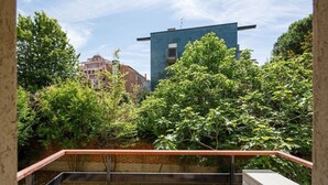 Blick von einem Balkon mit Metallgeländer auf einen Innenhof mit Bäumen und einem blauen Gebäude im Hintergrund.