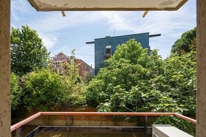 Blick von einem Balkon mit Metallgeländer auf einen Innenhof mit Bäumen und einem blauen Gebäude im Hintergrund.