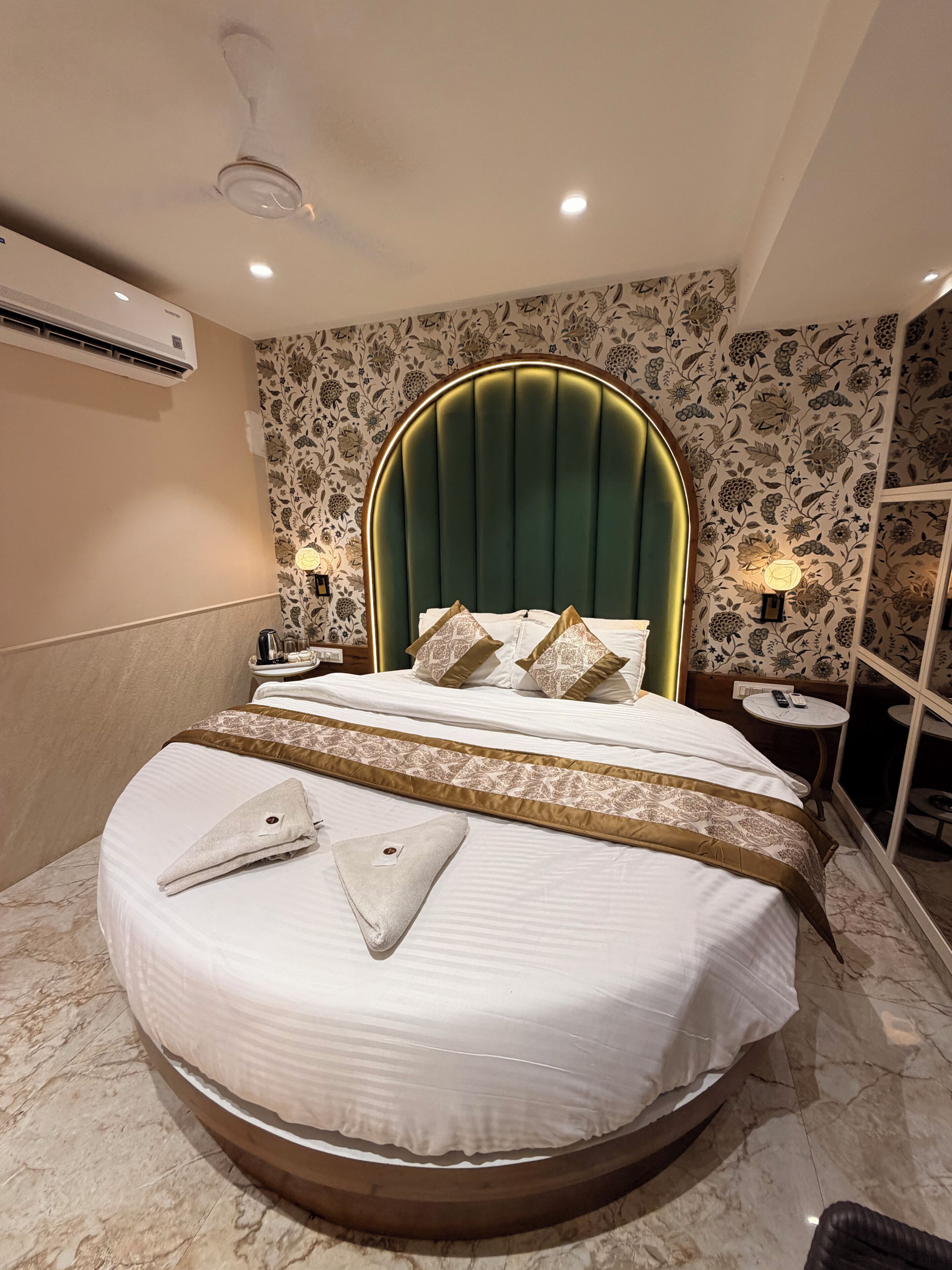 A hotel room with a round bed, a green upholstered headboard, floral wallpaper, a marble floor, and a ceiling fan.